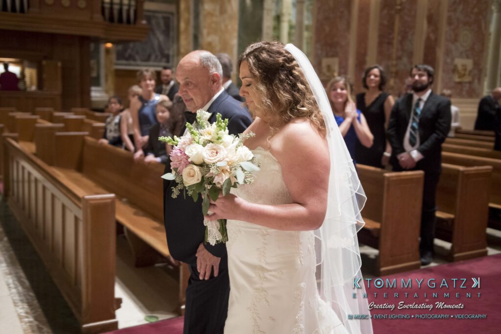 A bride holds a bouquet and walks down the aisle with an older man in a church, as guests watch.