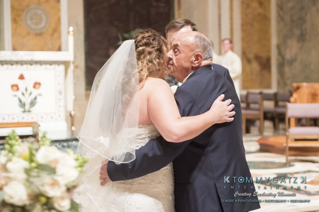 A bride in a white gown and veil hugs an older man in a suit inside a church. A religious altar is visible in the background.