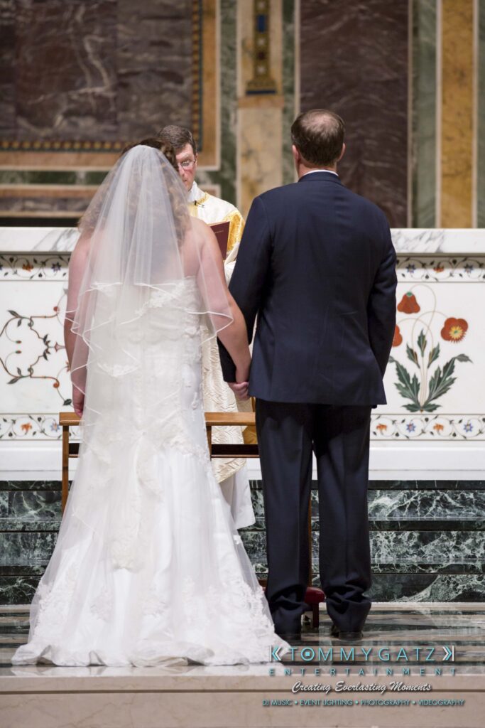 A bride and groom stand facing a priest at the altar during a wedding ceremony, with intricate designs on the wall behind them.