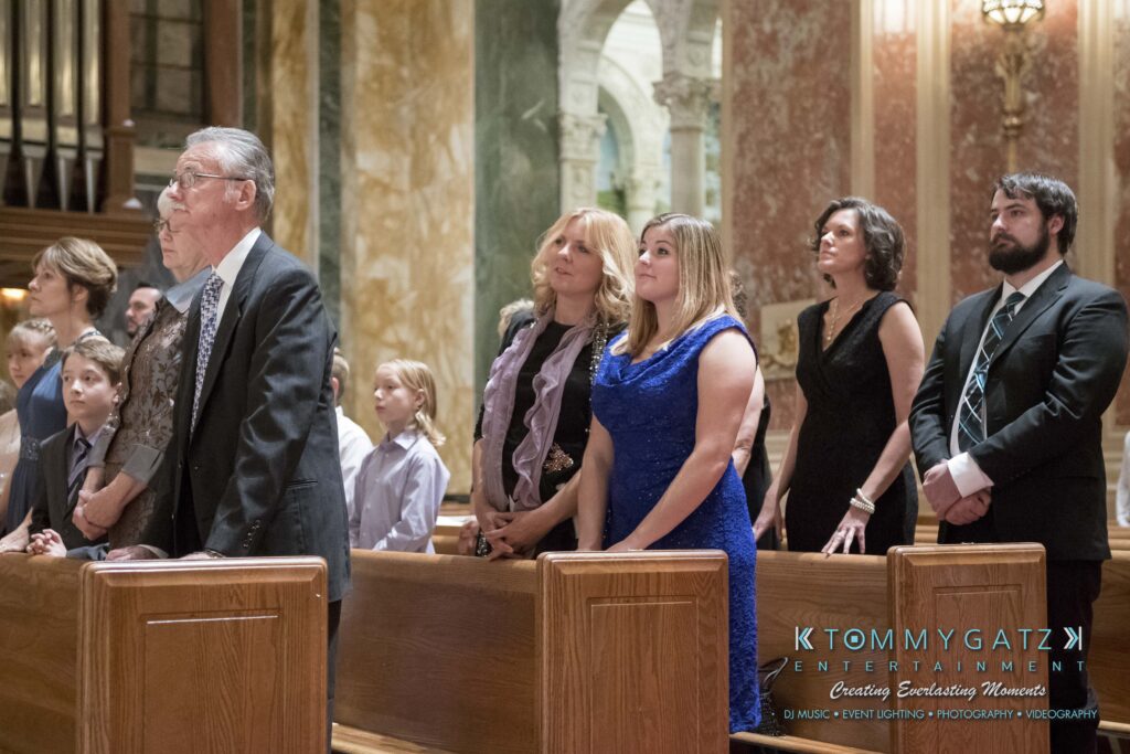 A group of people stand in a church pew during a service. They are dressed formally, with men in suits and women in dresses.