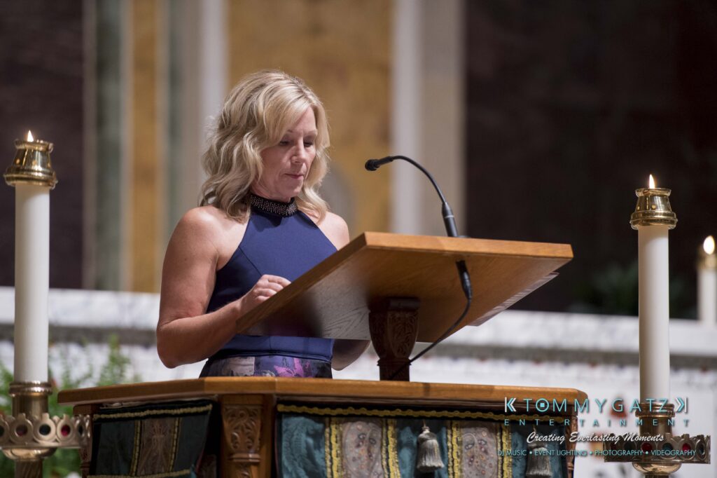 A woman in a blue dress stands at a lectern in a church, reading or speaking. Candles are visible on either side, and ornate decorations are in the background.