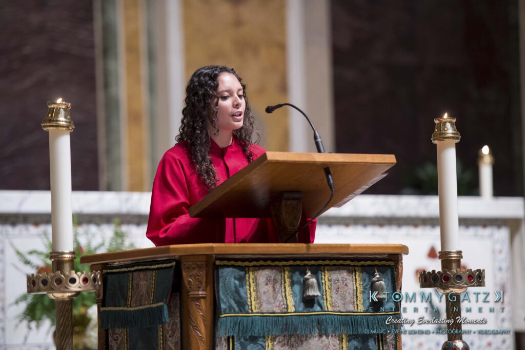A person in a red robe speaks at a wooden lectern with a microphone in a church setting. Candles are visible in the background.