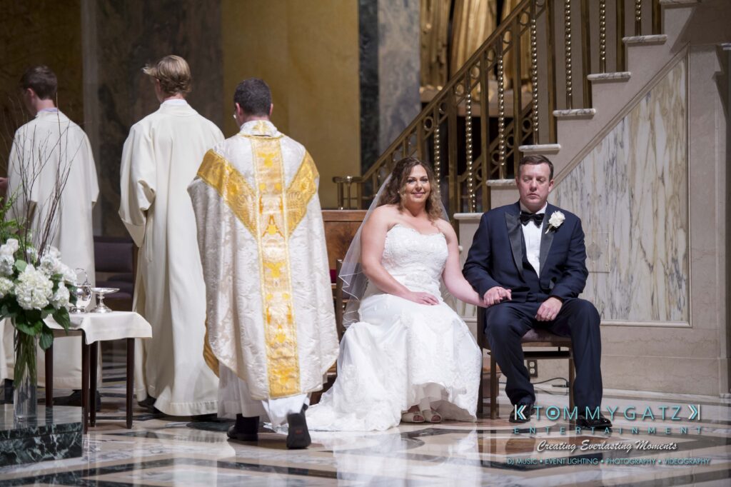 A bride and groom sit during a wedding ceremony in a church. A priest in a gold and white vestment stands nearby.