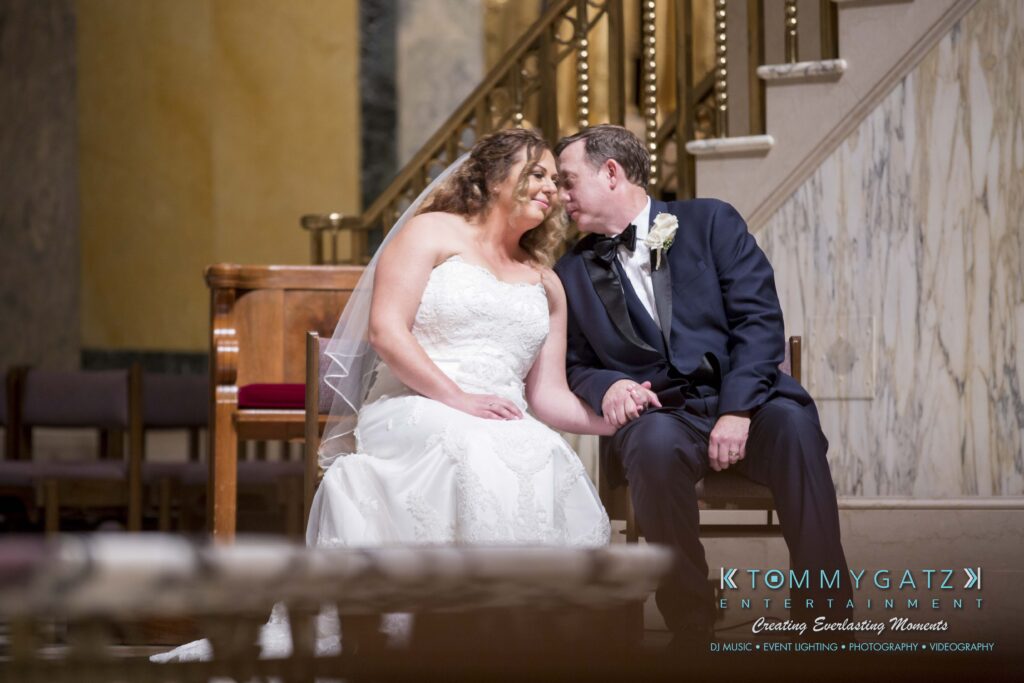 A bride and groom sit closely on a bench indoors, dressed in formal wedding attire.