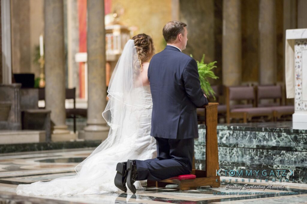 A bride and groom kneel on a bench in a church, facing the altar. The bride wears a white gown and veil, and the groom is in a dark suit.