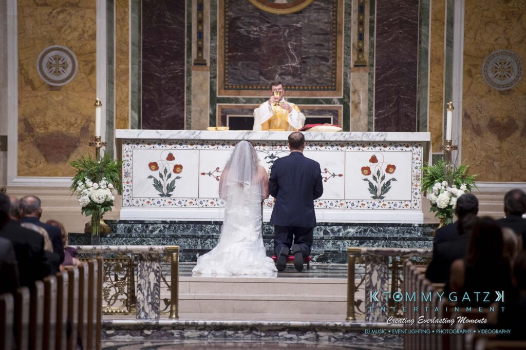 A couple kneels before a priest at an ornate altar during a wedding ceremony, with guests seated in the background.