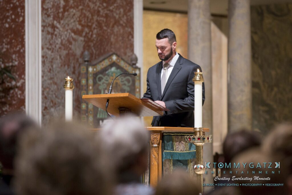 A man in a suit stands at a wooden podium in a church, reading from a paper. Candles and ornate decorations are visible in the background.