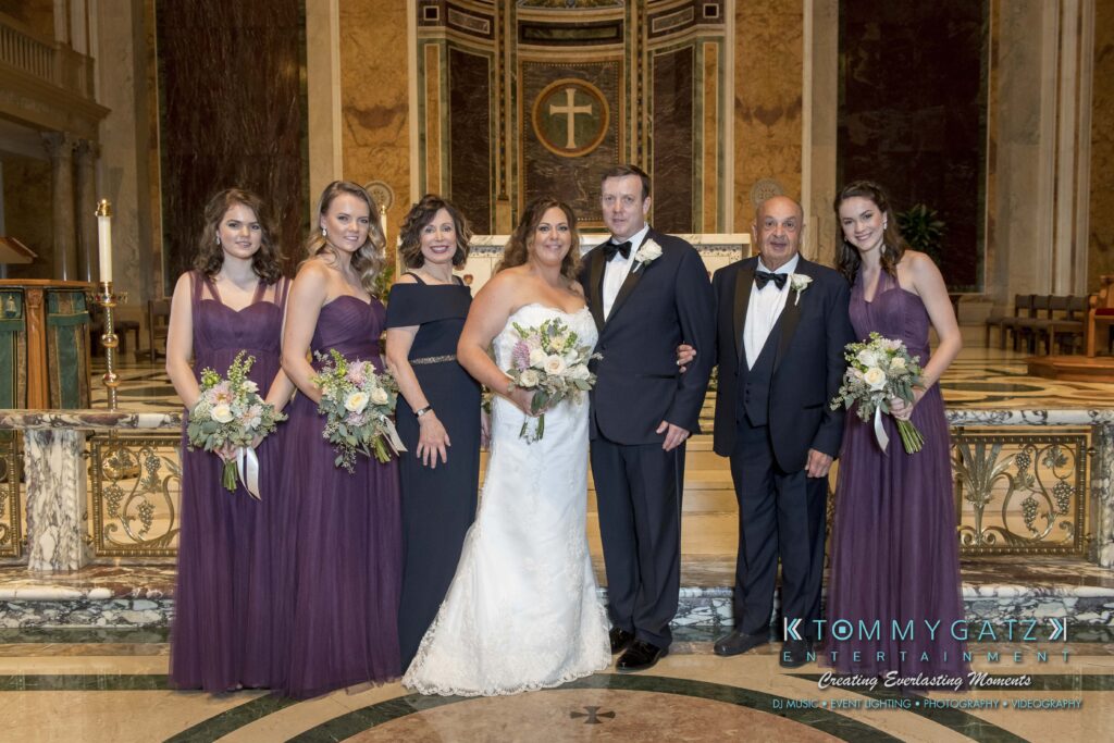A bride and groom stand with four women in purple dresses and an older man in a black suit, all posing in an ornate venue. The women hold bouquets.