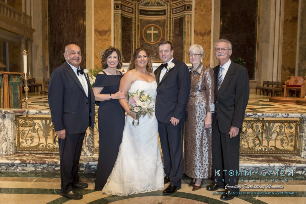 A bride and groom stand with four adults in formal attire in a decorated hall.