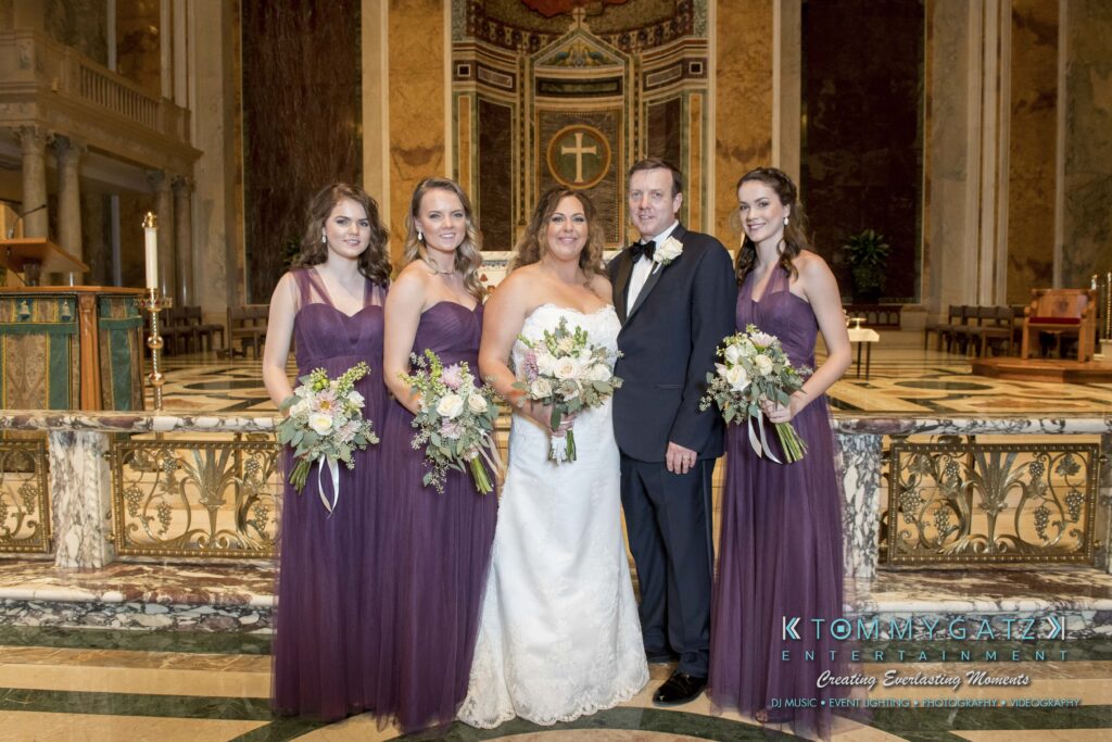 A wedding couple poses with four bridesmaids in purple dresses, each holding a bouquet, inside an ornate church setting.