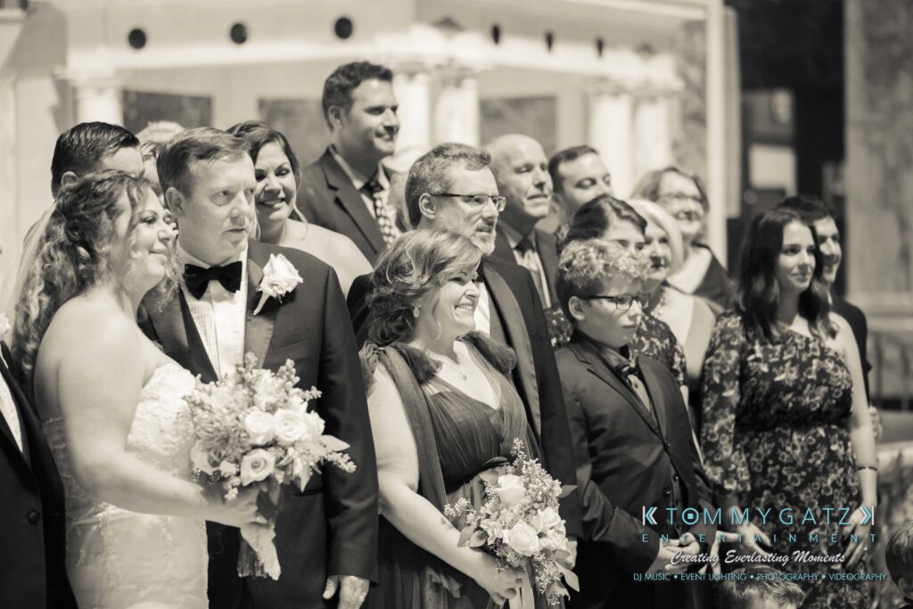 A black and white photo of a wedding group. The bride and groom stand in front, surrounded by guests. The bride holds a bouquet.