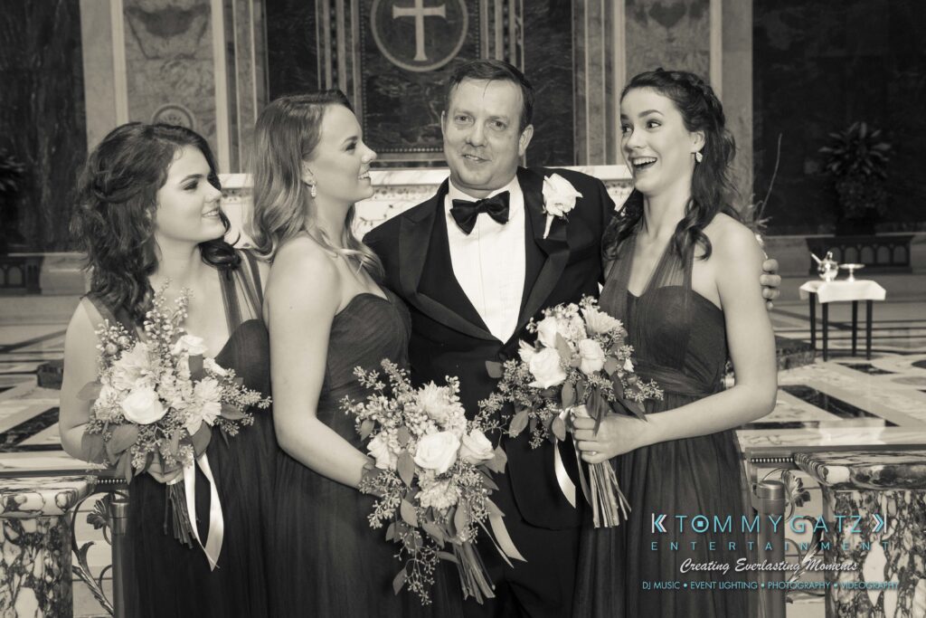 A man in a tuxedo stands with three women in dresses holding bouquets in a formal setting. They are smiling and looking at each other.