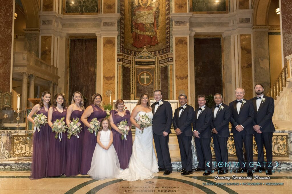 A wedding party stands in a church interior. The bride and groom are surrounded by bridesmaids in purple dresses and groomsmen in black suits. A young girl in a white dress stands in front.