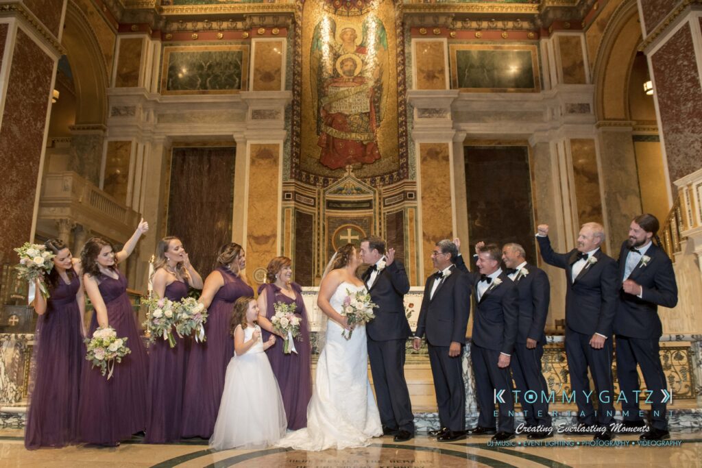 Wedding party celebrates as bride and groom kiss in an ornate church setting. Bridesmaids in purple dresses hold bouquets; groomsmen in suits cheer.