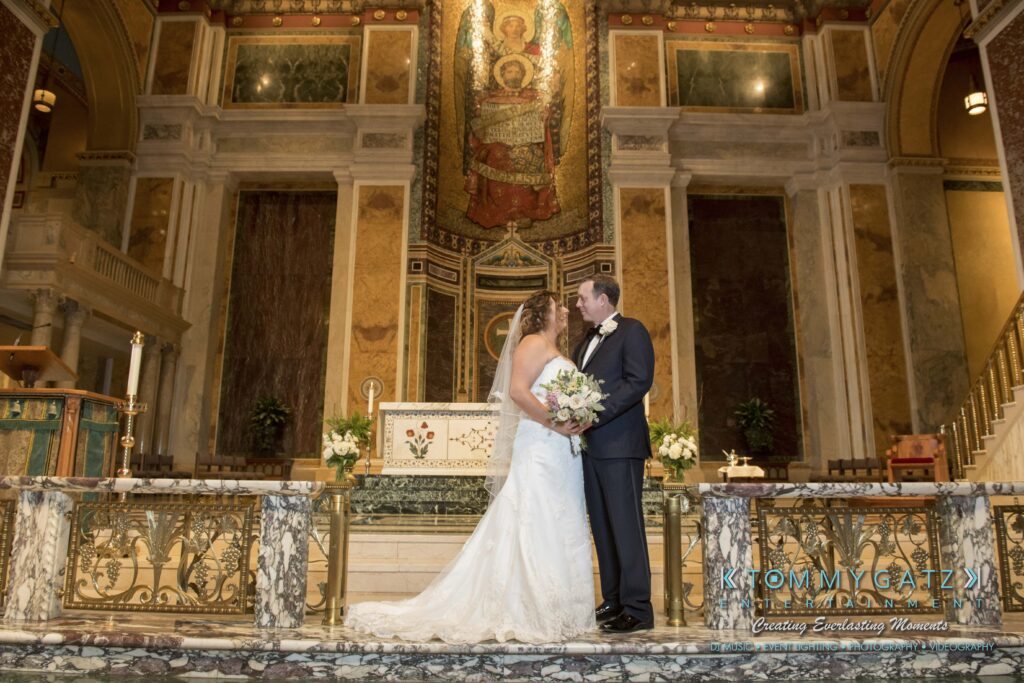A bride and groom kiss at the altar in an ornate church, holding a bouquet, with a religious mosaic in the background.