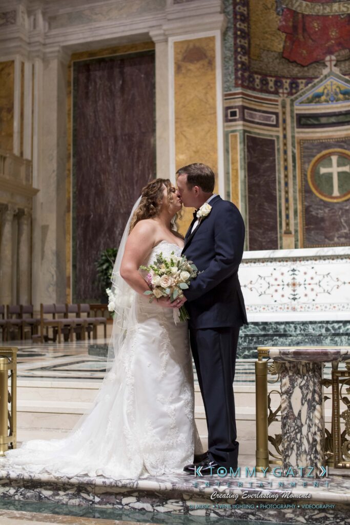 A bride and groom share a kiss at the altar in a church, with decorative backgrounds and floral arrangements.