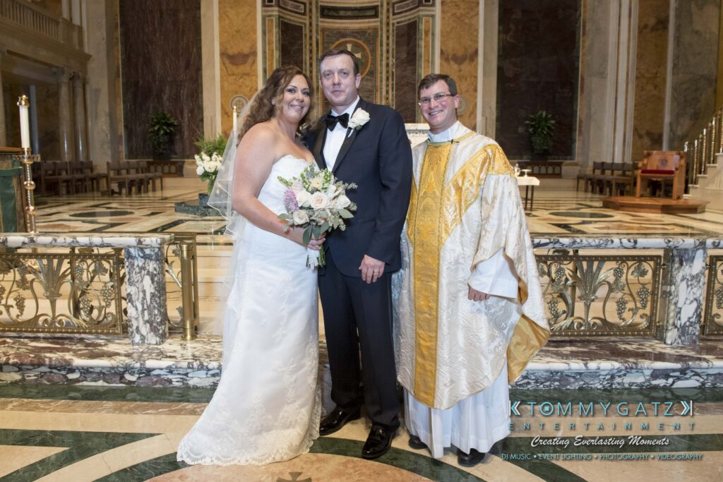 Bride and groom stand with a priest in a church, smiling for the photo. The bride holds a bouquet, and the priest wears golden vestments.