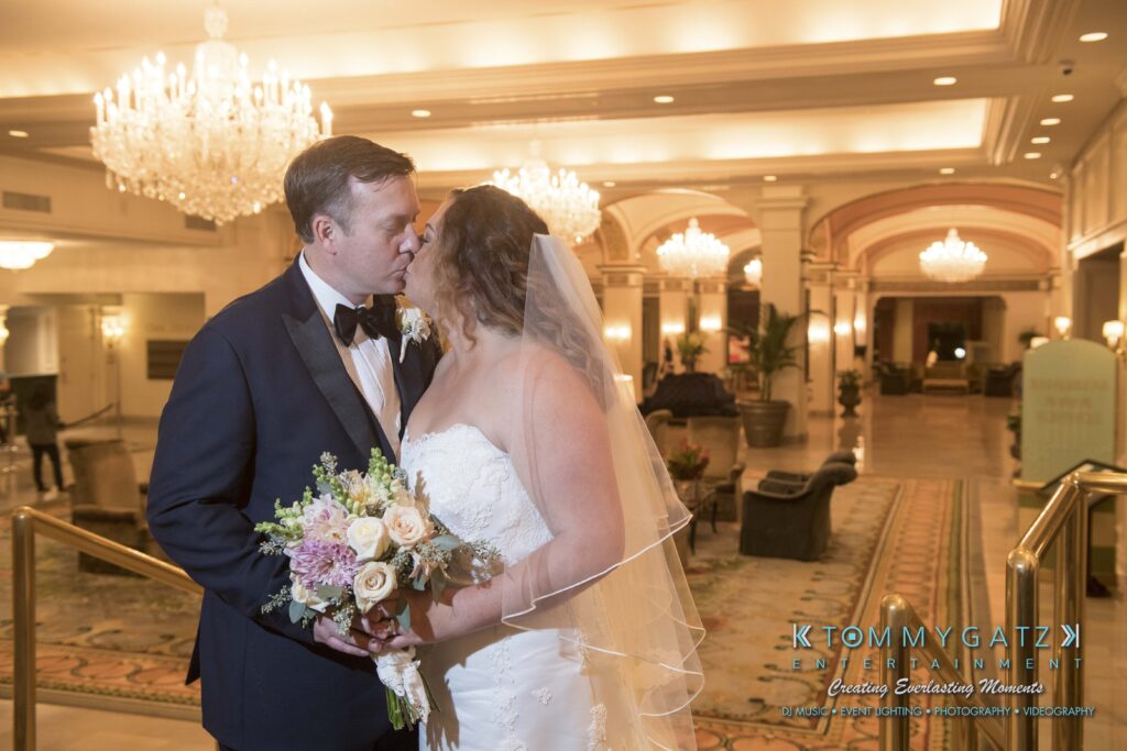 A bride and groom share an intimate moment in an elegant, chandelier-lit lobby. The bride holds a bouquet, and both are dressed in formal attire.