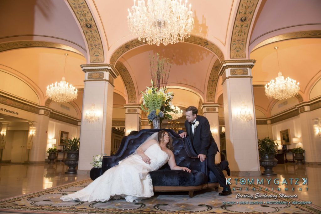 Bride and groom in formal attire pose together on a blue sofa in an ornate ballroom with chandeliers and floral arrangements.