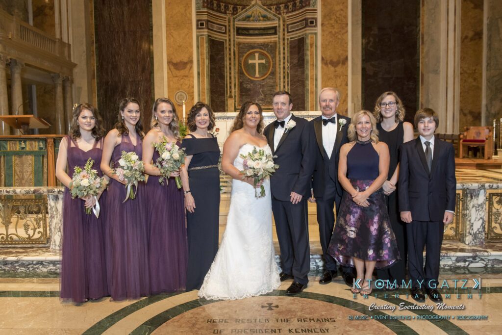 A bride and groom stand with family members in a church, posing for a group photo. The women wear purple dresses, and the men are in formal attire. The bride holds a bouquet.