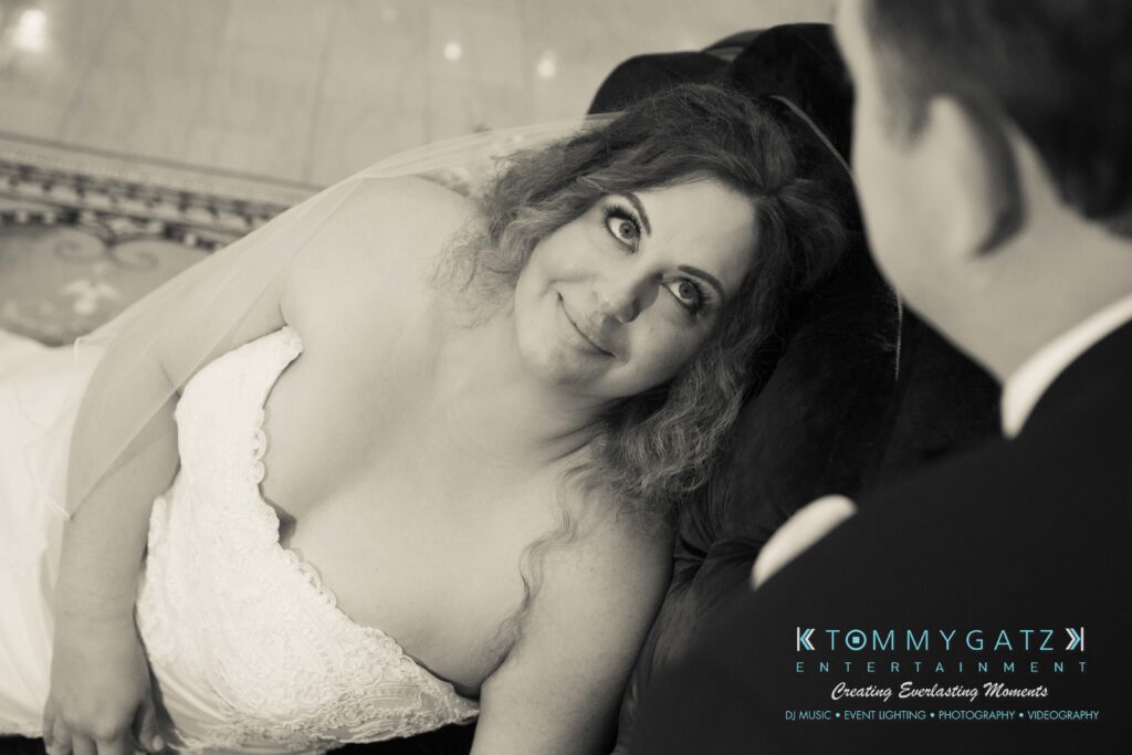 Bride in white dress and veil gazes upward smiling. Groom's back visible in foreground. Elegant setting with patterned floor.
