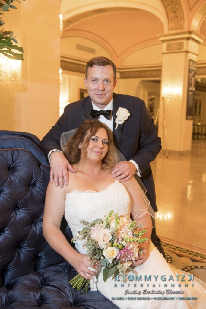 A bride in a white gown holds a bouquet while sitting on a sofa, with a groom in a tuxedo standing behind her. They are in an elegantly decorated indoor setting.