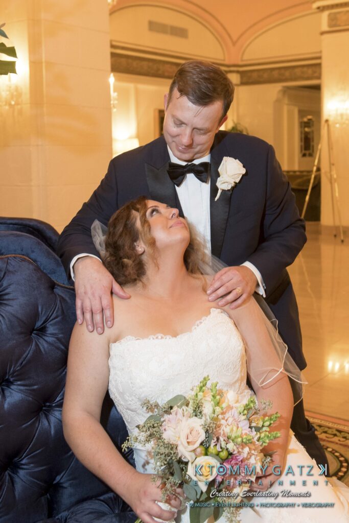 A bride and groom in wedding attire pose indoors. The bride holds a bouquet and sits, looking up at the groom, who stands behind with a hand on her shoulder.