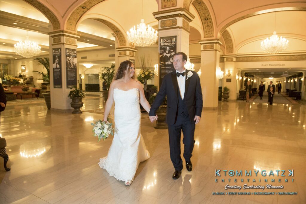 Bride in a white gown and groom in a black tuxedo walk hand in hand in an ornate hall with chandeliers and arched ceilings.