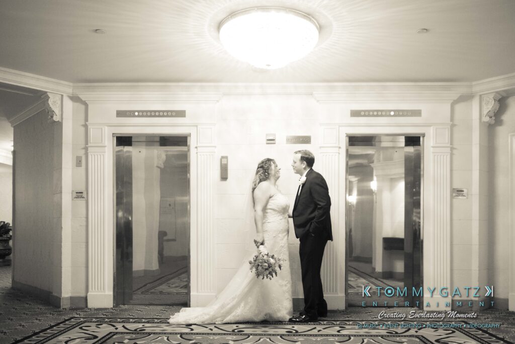 Bride and groom standing together in a hotel lobby, both smiling at each other. The bride holds a bouquet. The setting is elegant with a chandelier above and elevators behind them.