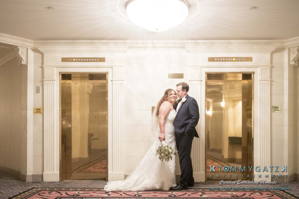 A bride and groom stand affectionately together in front of elevators. The bride holds a bouquet, and both are dressed in wedding attire.