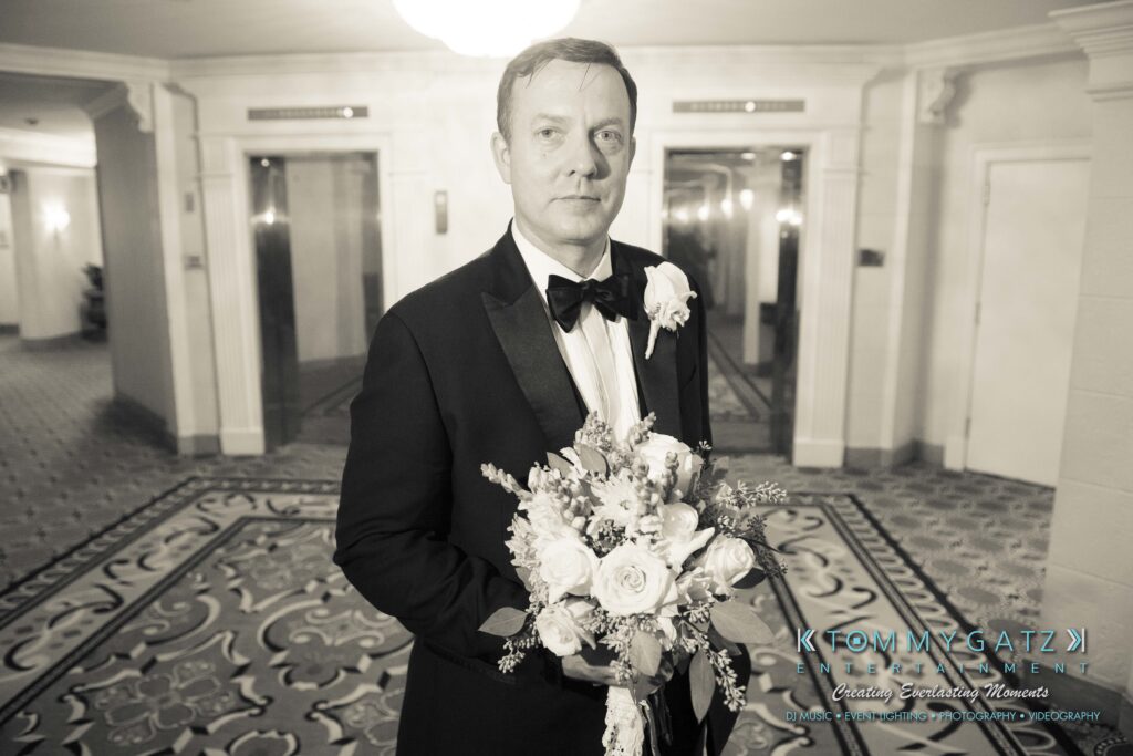 Man in a tuxedo holds a floral bouquet in a hotel lobby.