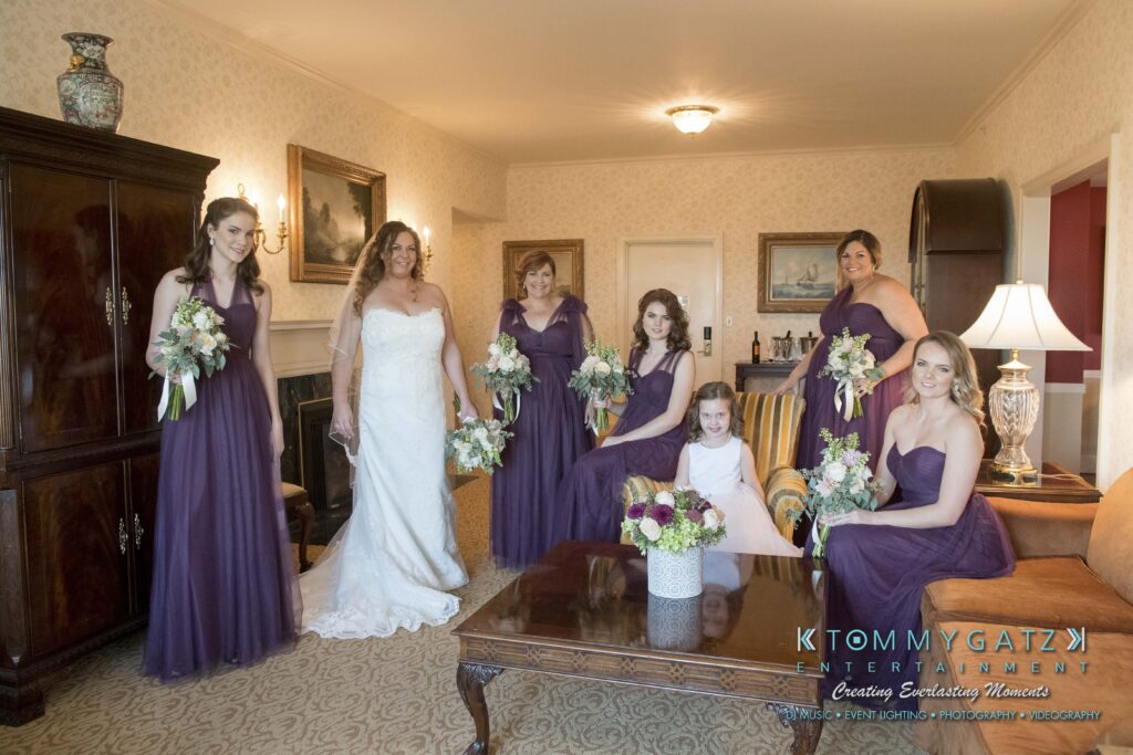 Bride with five bridesmaids in purple dresses and a young girl in white, posing in a warmly furnished room. The bridesmaids hold flower bouquets.