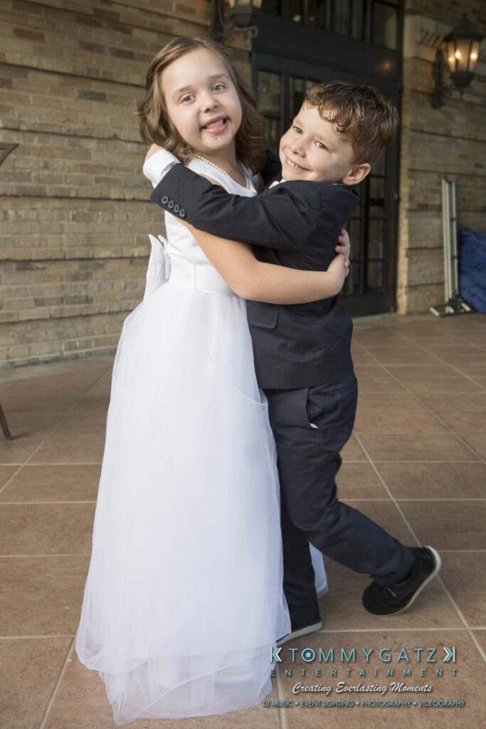 Two children dressed formally, a girl in a white dress and a boy in a suit, joyfully hugging and smiling on a tiled floor with a brick wall background.