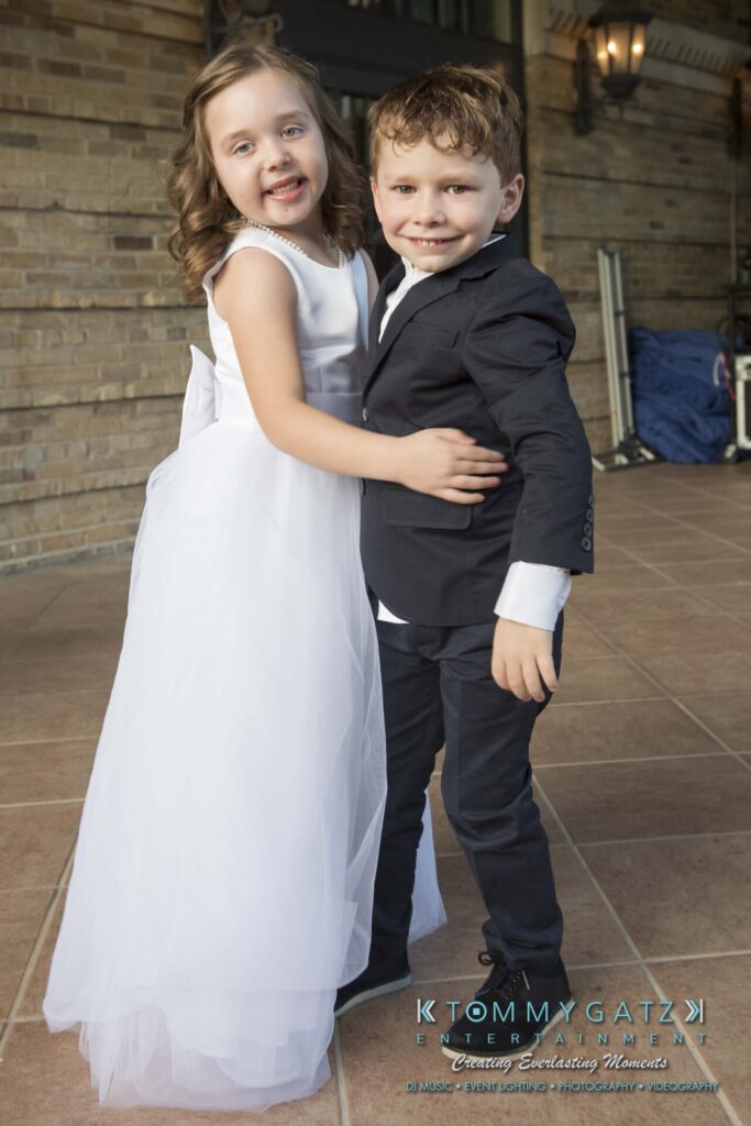 A young girl in a white dress and a young boy in a dark suit smile and pose together in front of a brick wall.