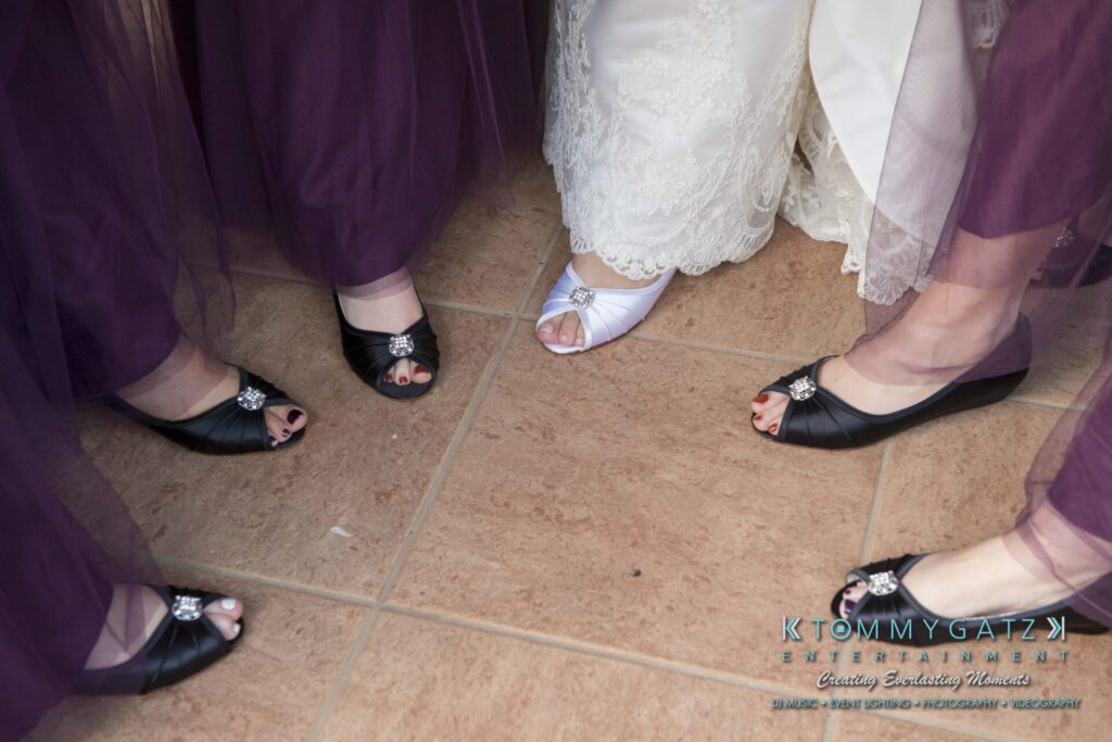 Bridal party in purple dresses, standing in a circle, showing their black shoes with rhinestone accents around a bride wearing white shoes.