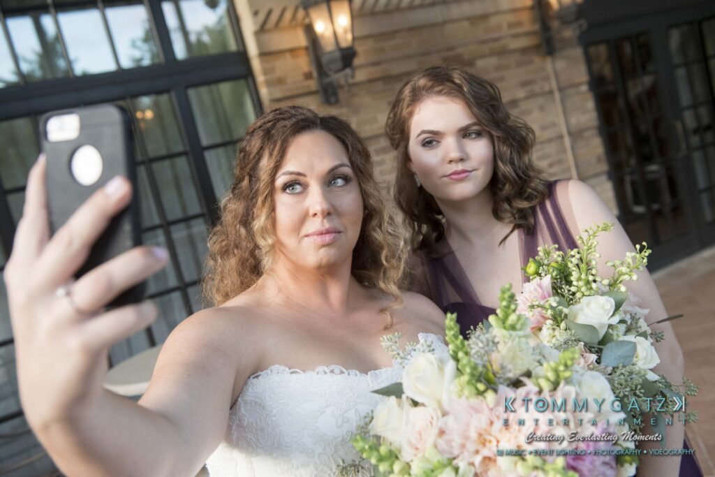 Two women taking a selfie; one in a white dress holding a bouquet, the other in a purple dress, standing in front of a brick building.