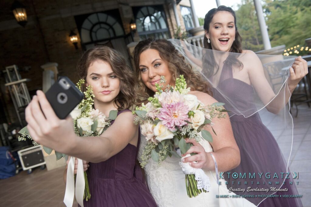 Three women in dresses take a selfie at a wedding. The bride is in the center holding a bouquet.