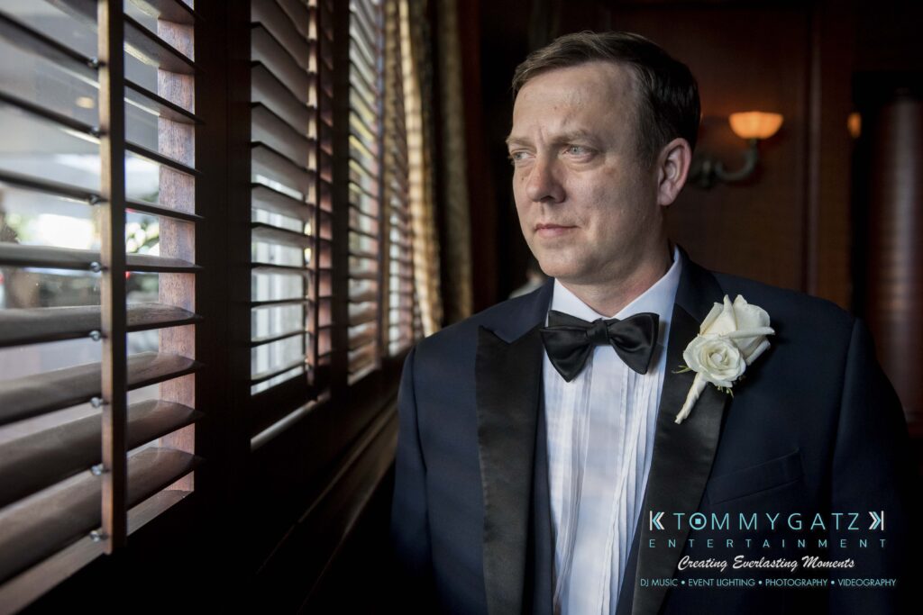 Man wearing a tuxedo with a white rose boutonniere, looking out a window with blinds, in a dimly lit room.