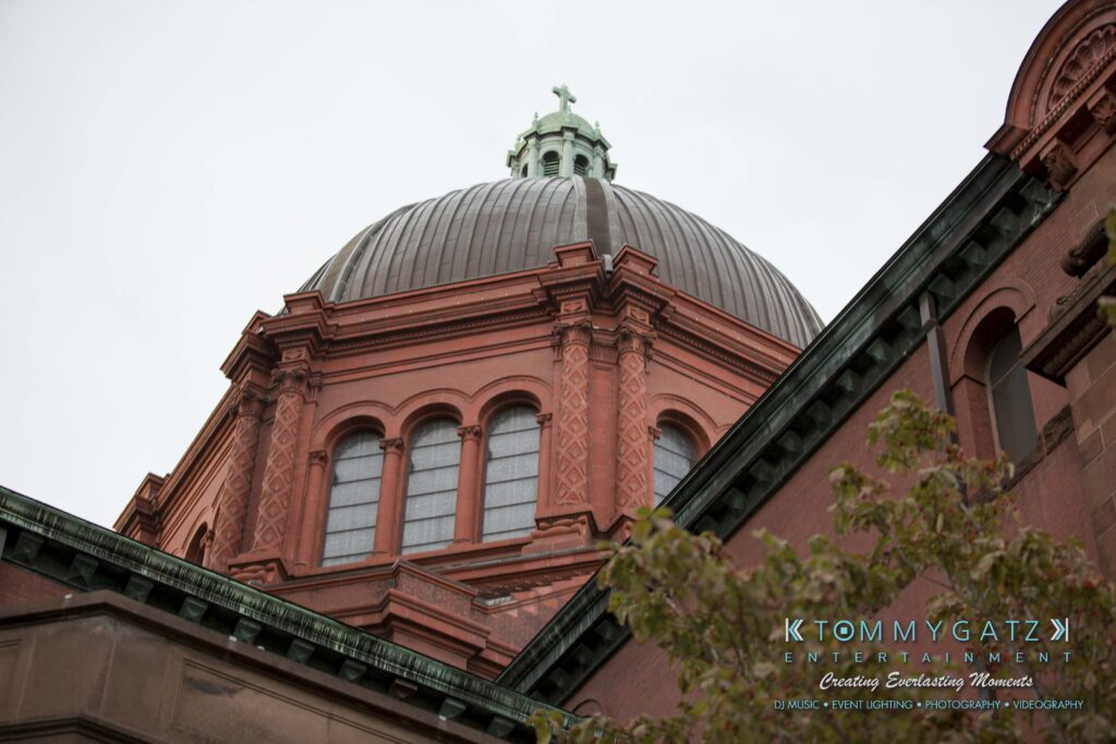 Close-up view of a large, red-brick dome with arched windows, set against a cloudy sky. Green leafy branches are partially visible in the foreground.