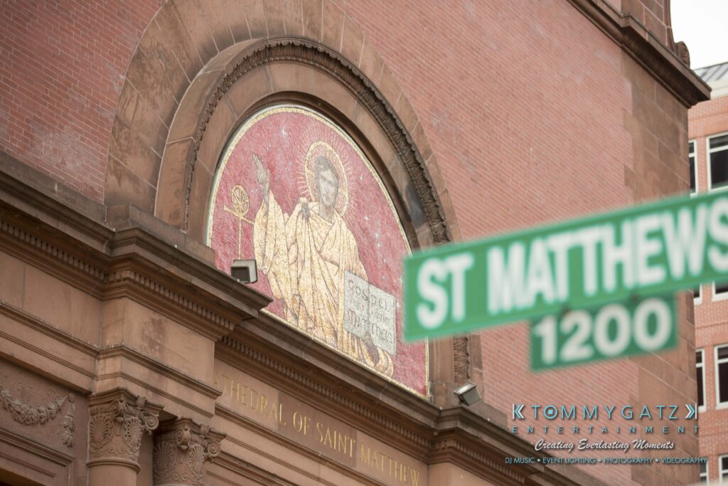 Mosaic of a religious figure on a red brick building, labeled "CATHEDRAL OF SAINT MATTHEW THE APOSTLE." A green street sign with "ST MATTHEW'S 1200" is in the foreground.