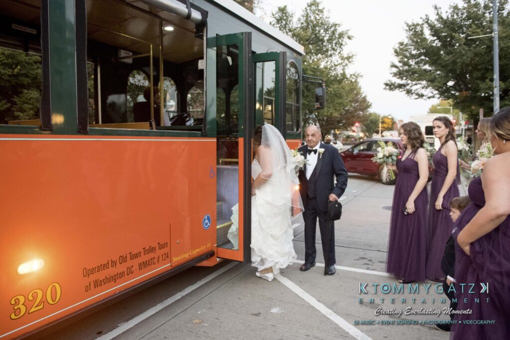 A bride and a man in formal wear prepare to board an orange bus, with bridesmaids in purple dresses nearby.