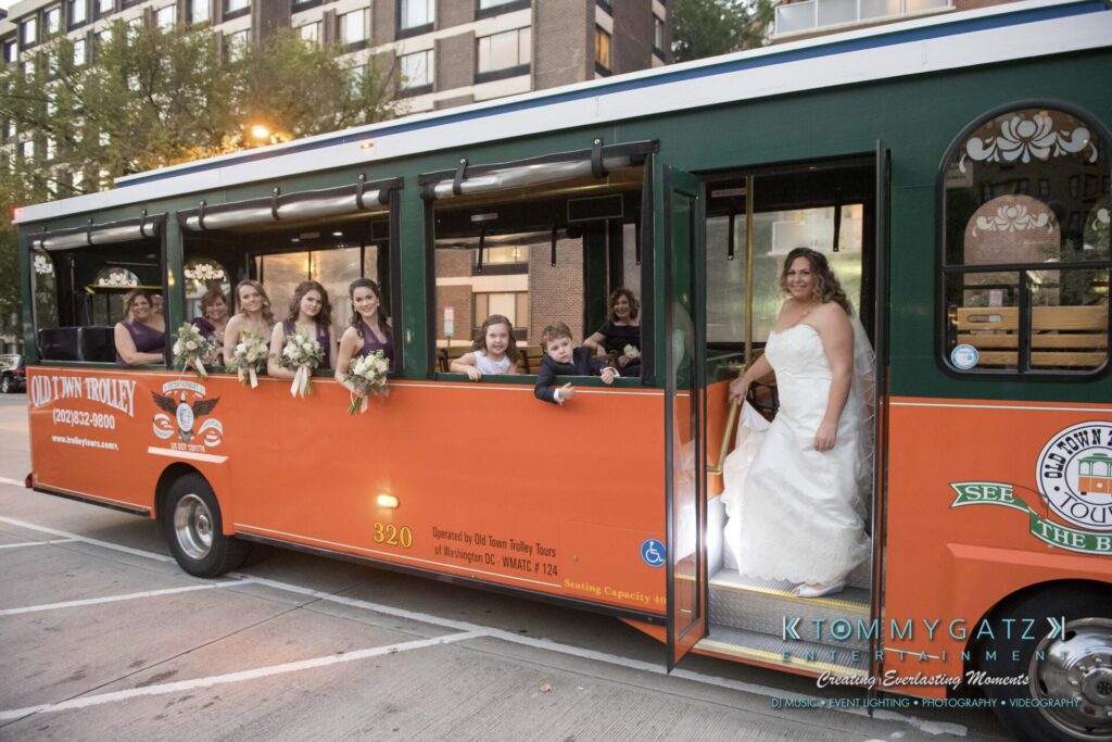 A bride in a white gown stands at the steps of an orange trolley bus, while the bridal party, holding bouquets, is seated inside.