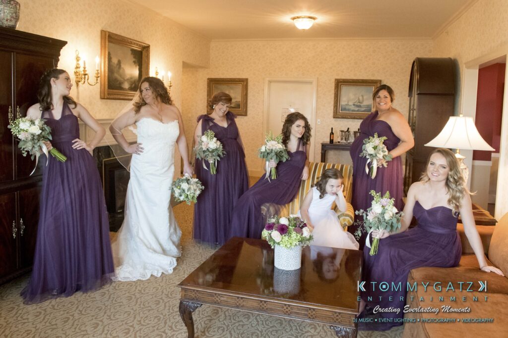 Bride in white gown with bridesmaids in purple dresses holding bouquets, posed in a room with elegant decor.