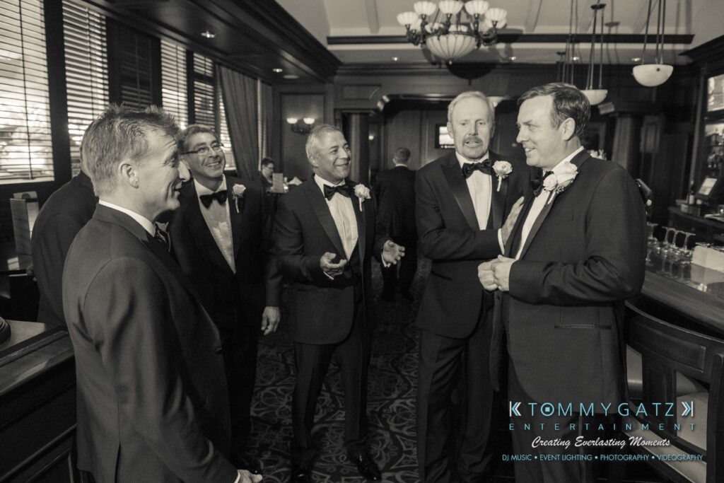 Five men in formal suits stand and converse in an elegant room with wood paneling and chandeliers.