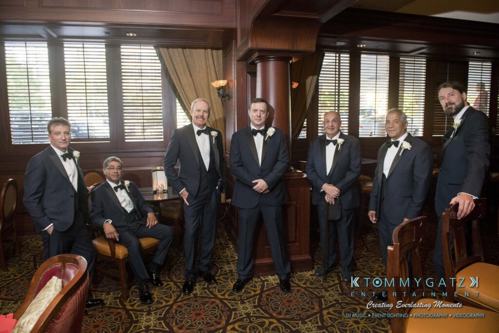 Seven men in formal suits pose in a wood-paneled room with striped windows and patterned carpet.