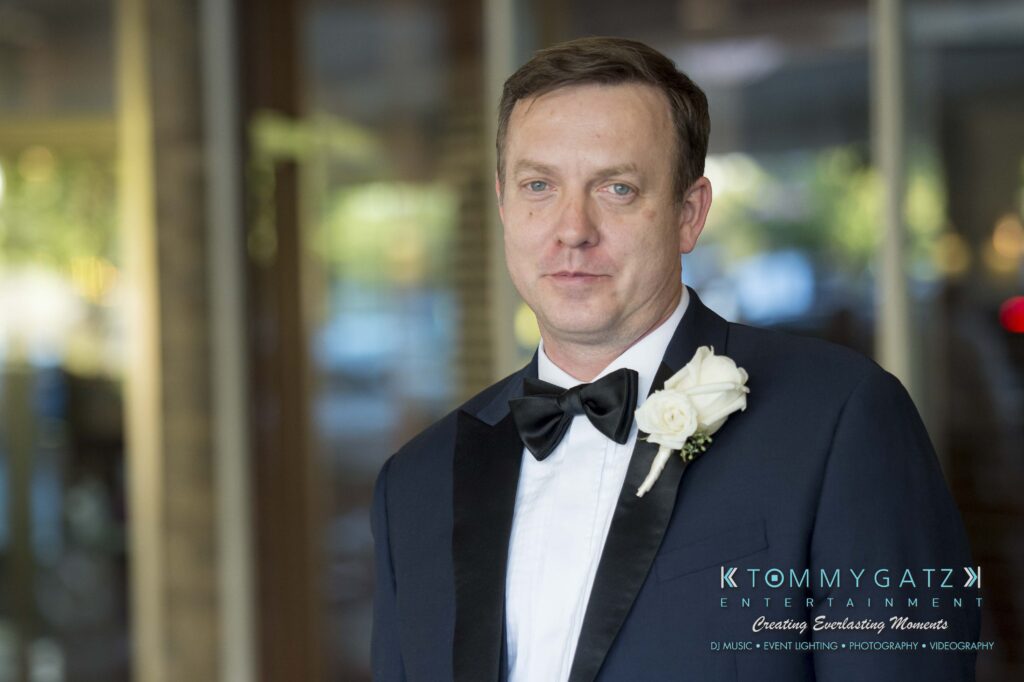 A man in a suit with a bow tie and floral boutonniere stands indoors, with a blurred background of lights.