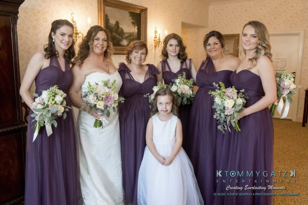 A bride in a white dress and six bridesmaids in matching purple dresses pose indoors, holding bouquets. A young girl in a white dress stands in front.