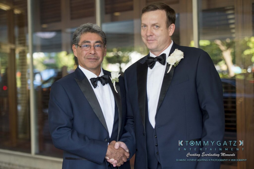 Two men in black tuxedos and bow ties shake hands, both wearing white boutonnieres. They stand indoors with glass and wooden paneling behind them.