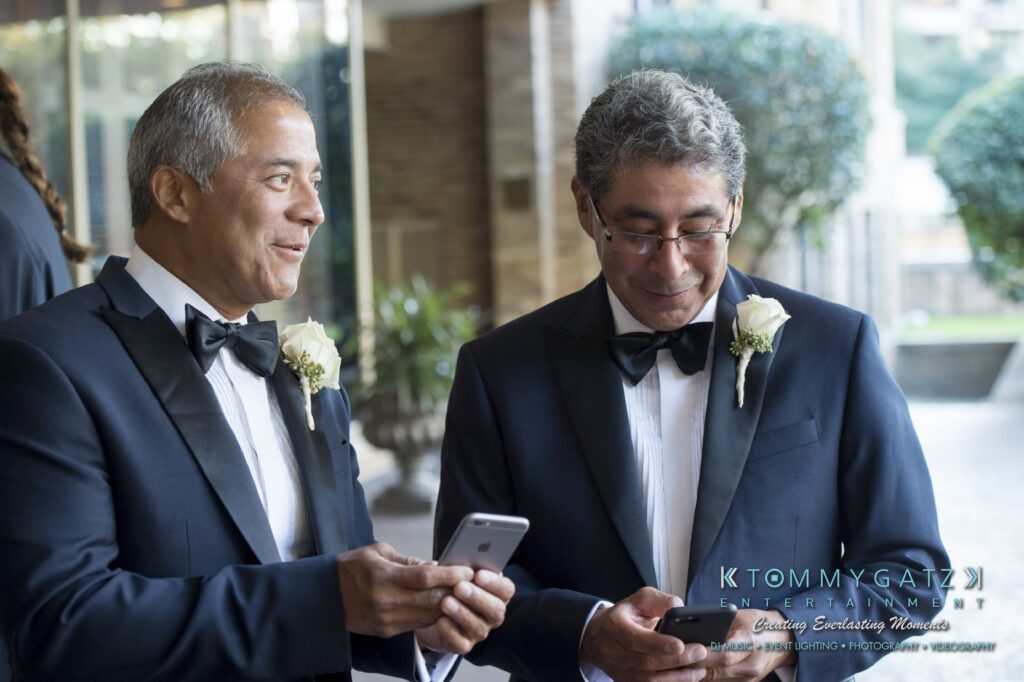 Two men in tuxedos stand next to each other, both looking at their phones. They are wearing white boutonnieres.