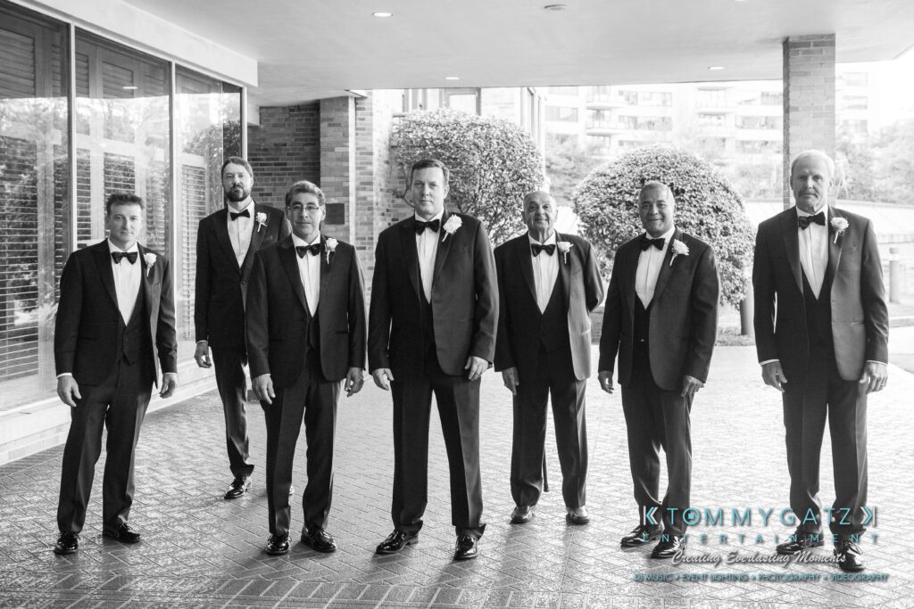 Seven men in tuxedos stand on a paved area outside a building with glass windows and shrubs, captured in black and white.